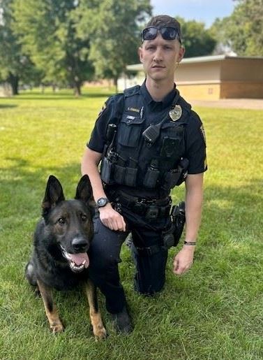 german shepard dog laying down with police officer kneeling next to him on the grass